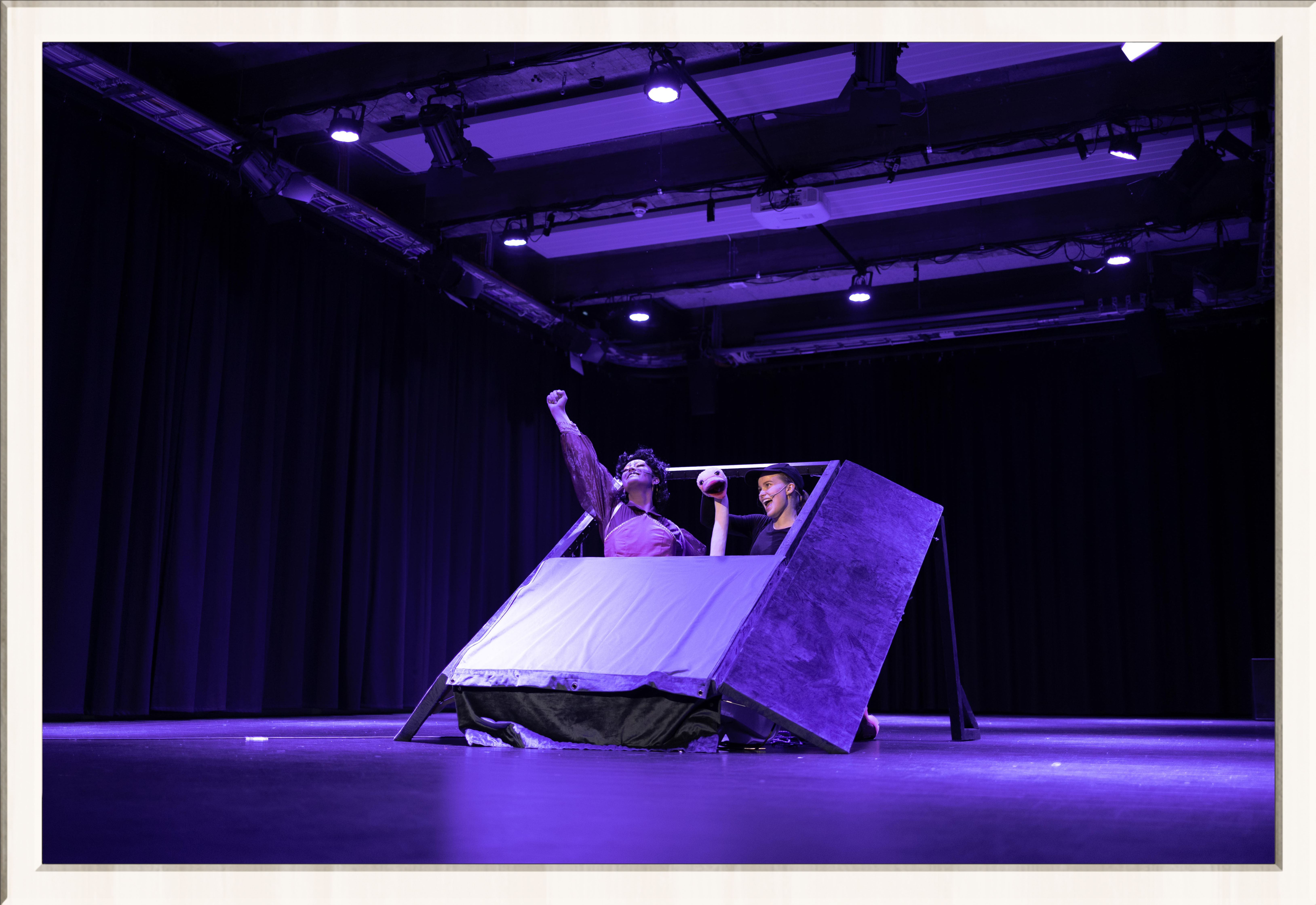 Two performers in a purple lit stage, one wearing a drag costume, the other an ostrich costume, performing a musical play for kids, framed in a thin white frame