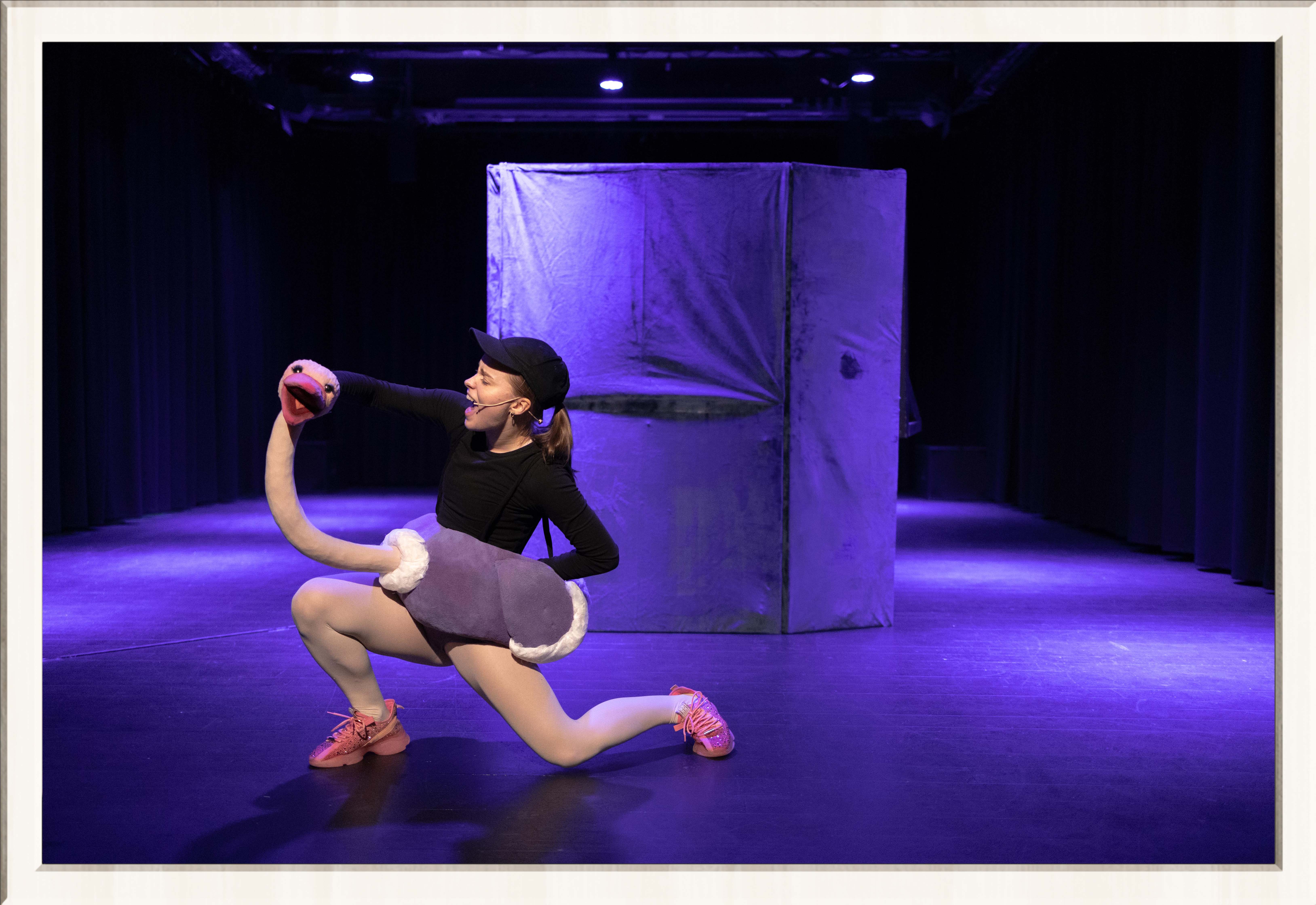 Two performers in a purple lit stage, one wearing a drag costume, the other an ostrich costume, performing a musical play for kids, framed in a thin white frame